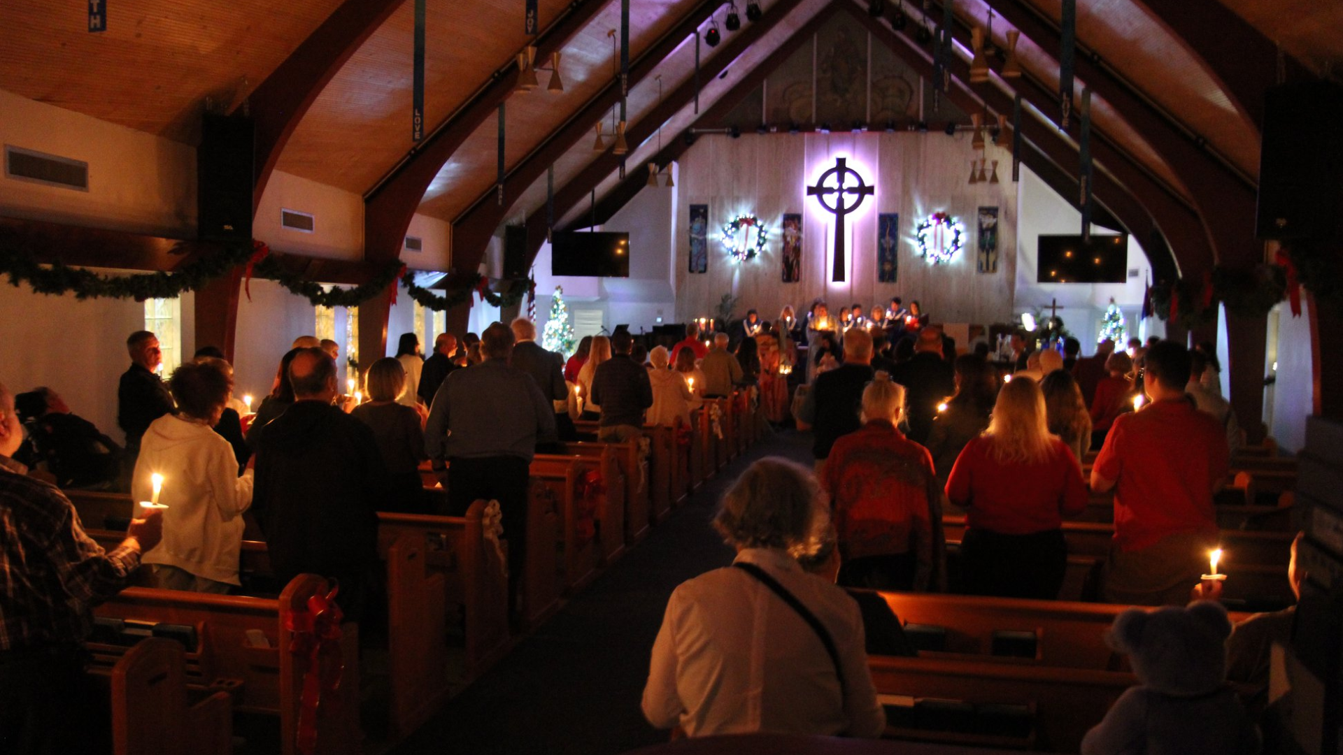 People holding candles in a dimly lit church during a service, cross illuminated on the altar.