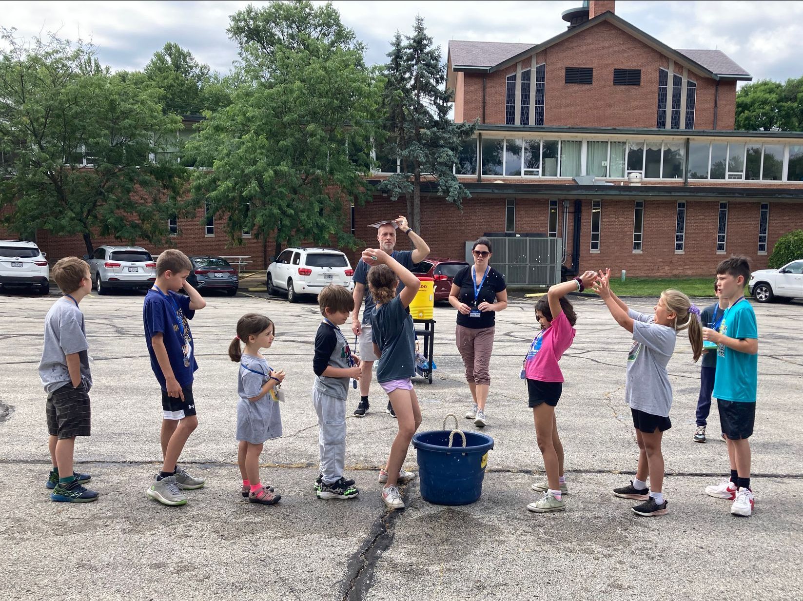 A row of children playing a game where they pass a water balloon over their heads in the church parking lot