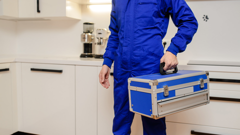 Person in blue overalls holding a blue and silver toolbox in a white kitchen.