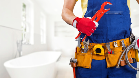 Plumber in blue overalls and tool belt holds red pliers in a bathroom setting.