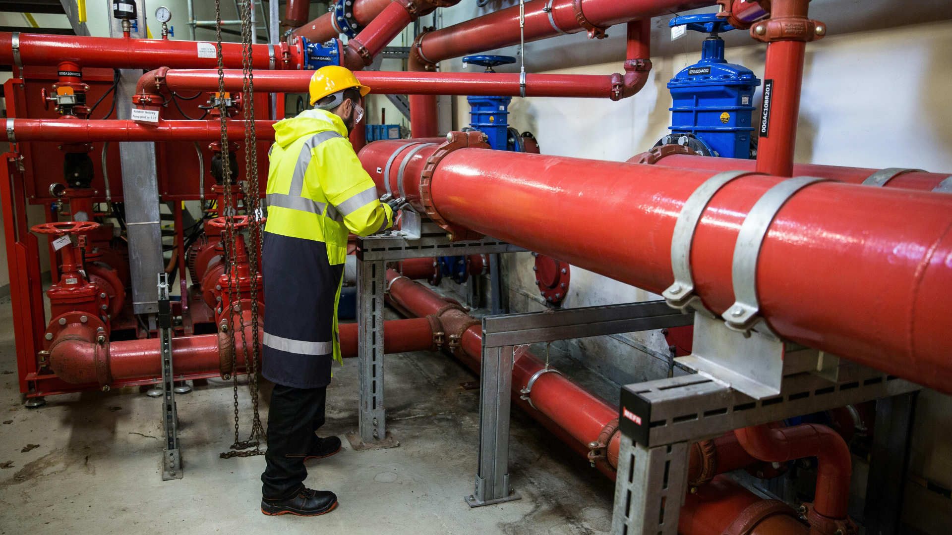 Worker in a yellow safety vest inspects red pipes in a mechanical room.