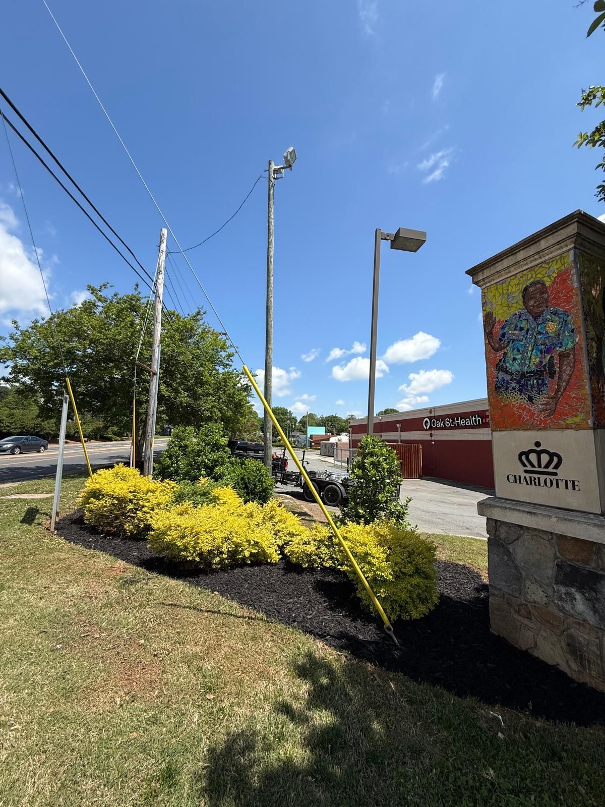 Entrance sign with mosaic art and landscaping, power lines, and streetlights on a sunny day.