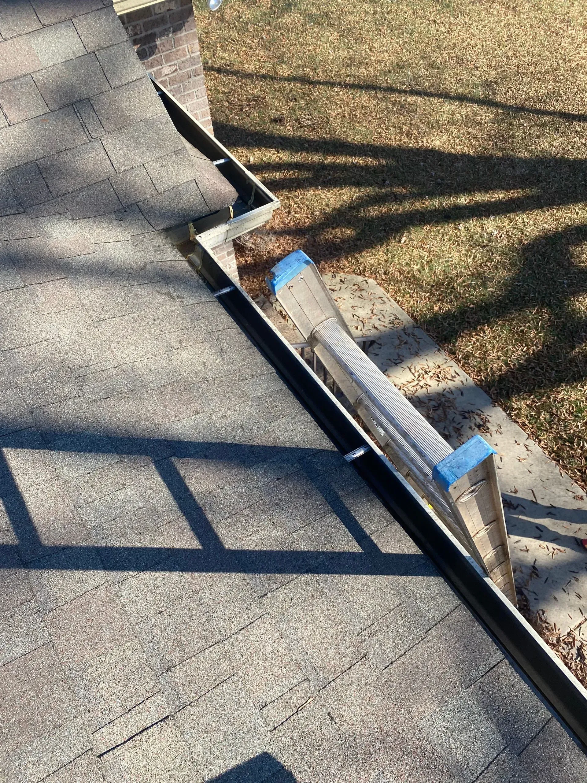Overhead view of a roof edge with a gutter partially filled with leaves. Shadow from a ladder present.