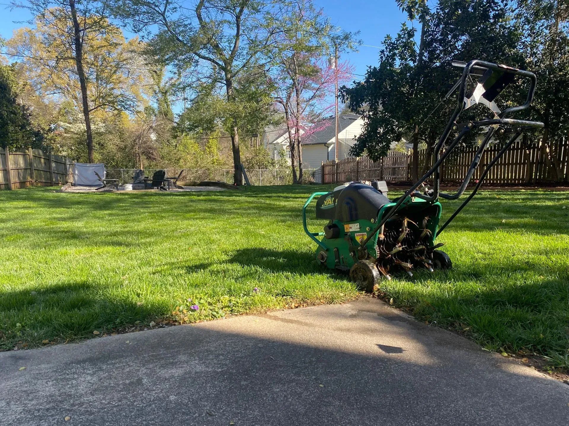 Lawn aerator on green grass near a paved driveway, surrounded by trees and a fence.