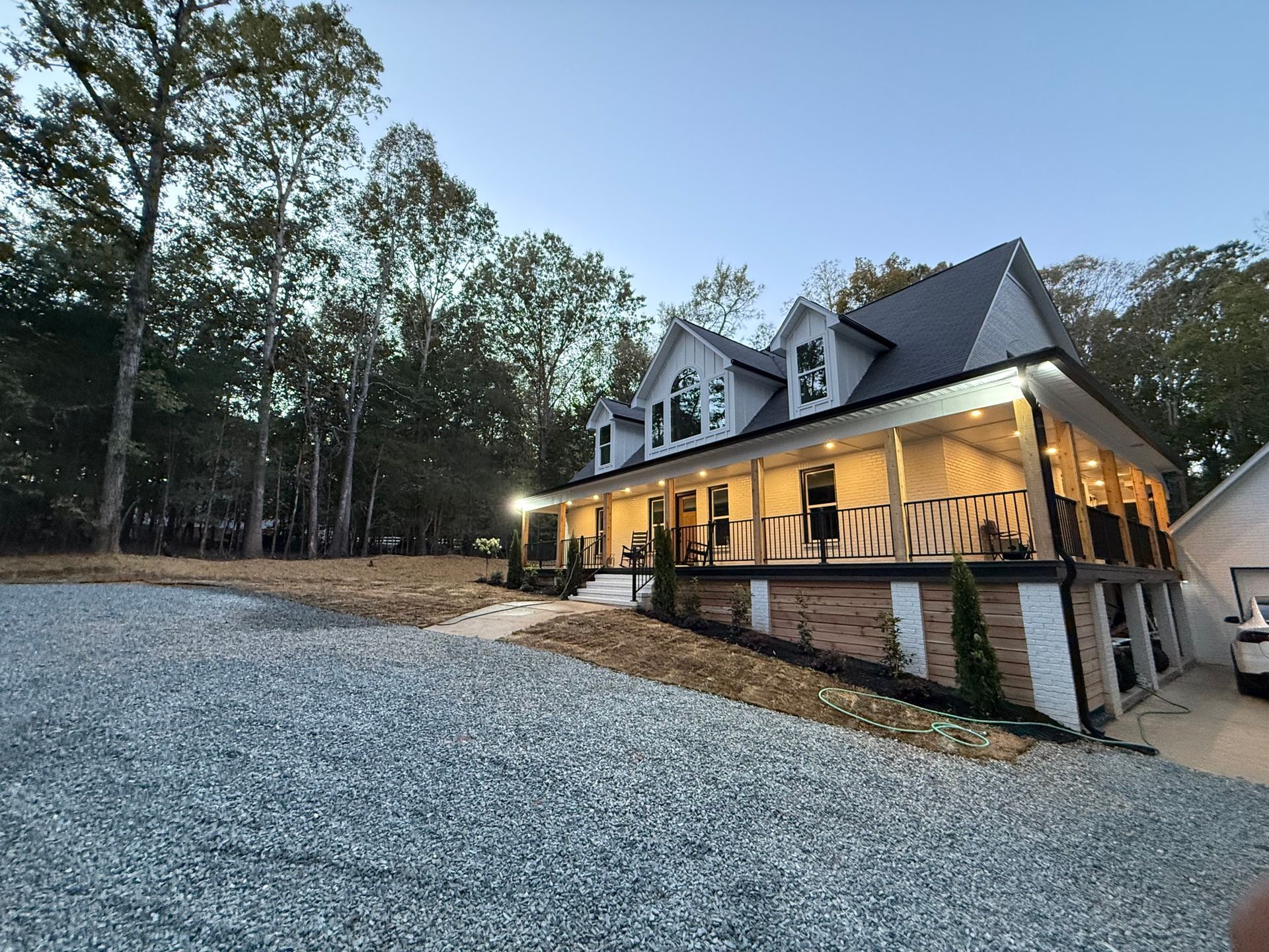 Two-story house with a large porch and gravel driveway surrounded by trees Its a Grading Job.