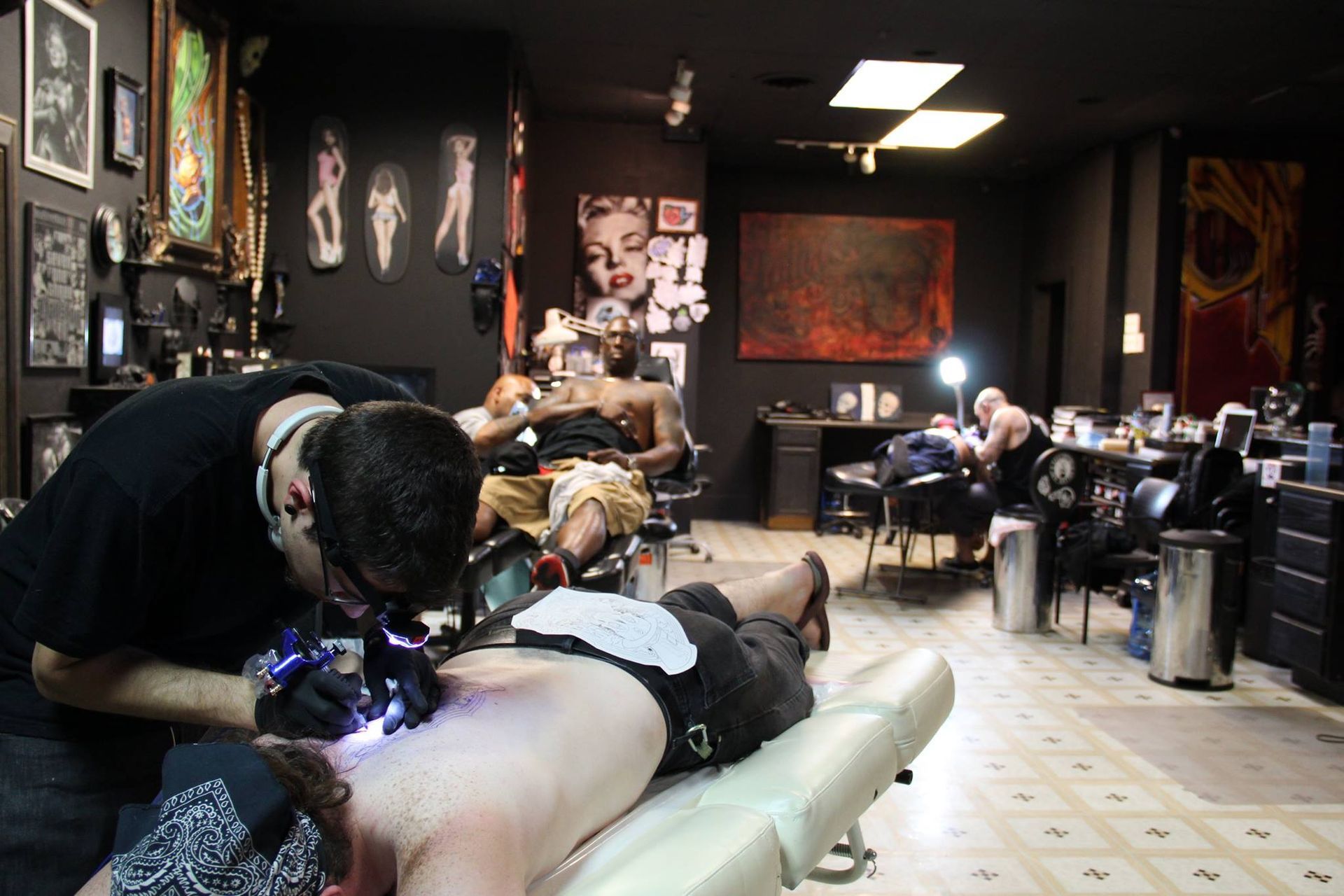 Tattoo artist working on client's back in a dimly lit tattoo parlor. Other customers present.