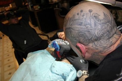 Person with head tattoo, tattoos a person's neck. Another person relaxes in the background.