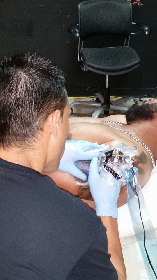 Tattoo artist at work. A person's back exposed; artist wearing gloves, leaning in with tattoo machine.