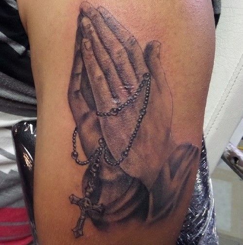 Tattoo of praying hands holding a rosary with a cross; black and white shading on arm.