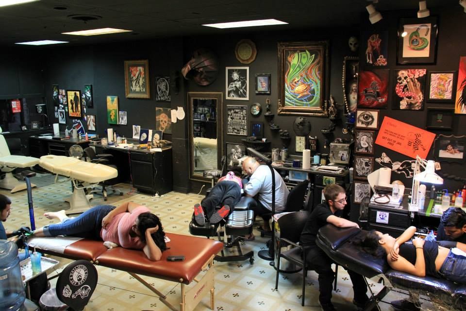 Interior of a tattoo parlor. People getting tattoos, artists working. Black walls, artwork, bright lights.