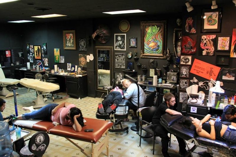 Interior of a tattoo parlor. People getting tattoos, artists working. Black walls, artwork, bright lights.