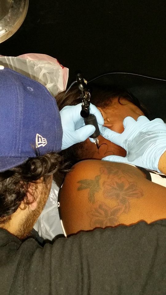 Person getting face tattoo, tattoo artist in gloves, visible tattoo on shoulder.