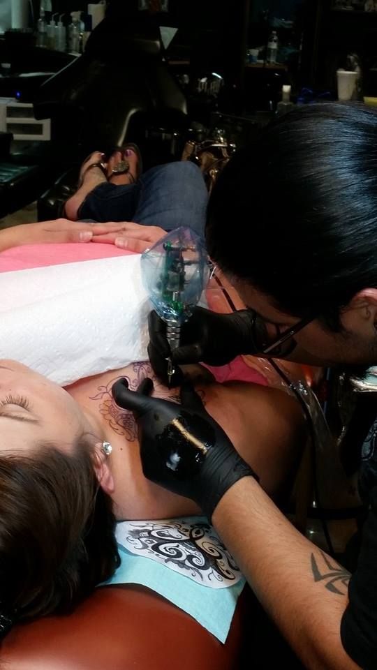 Tattoo artist in black gloves inks a design on a person's chest. Someone rests on a table.