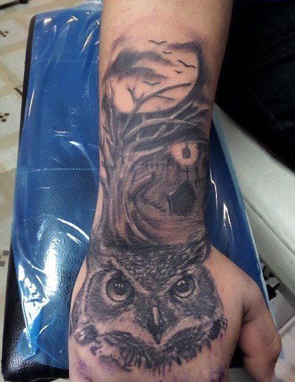 Tattoo of an owl on a wrist and hand. Features a dark owl with a moon, trees, and branches in gray and black.