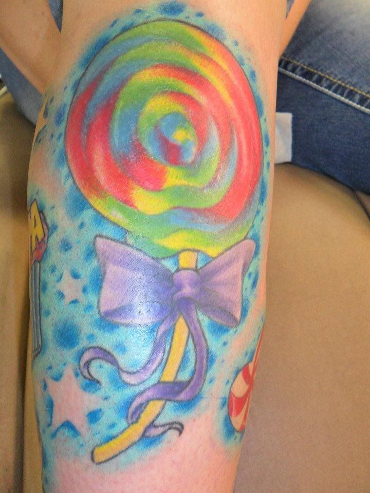Colorful lollipop tattoo on a leg, with a purple bow, stars, and blue background.