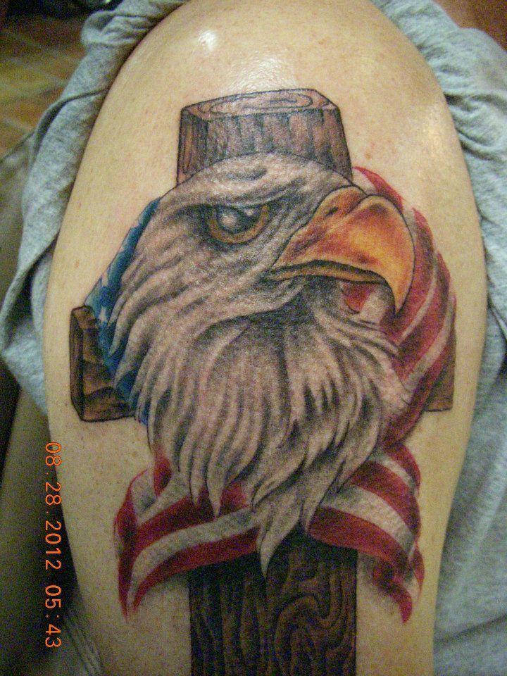 Tattoo of an eagle head, American flag, and cross on a shoulder. Eagle has a stern look.