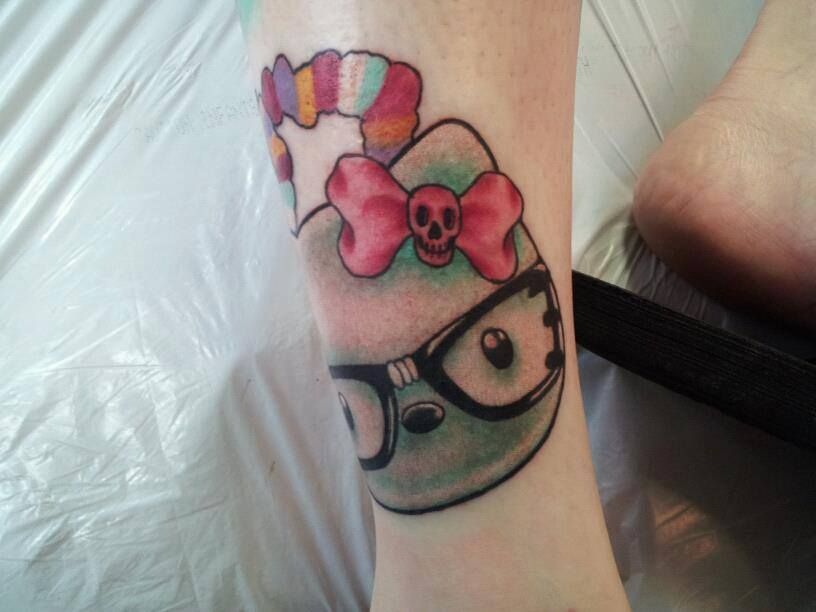 Tattoo of a green cartoon character wearing glasses and a pink bow with a skull, on a leg. Rainbow hair.