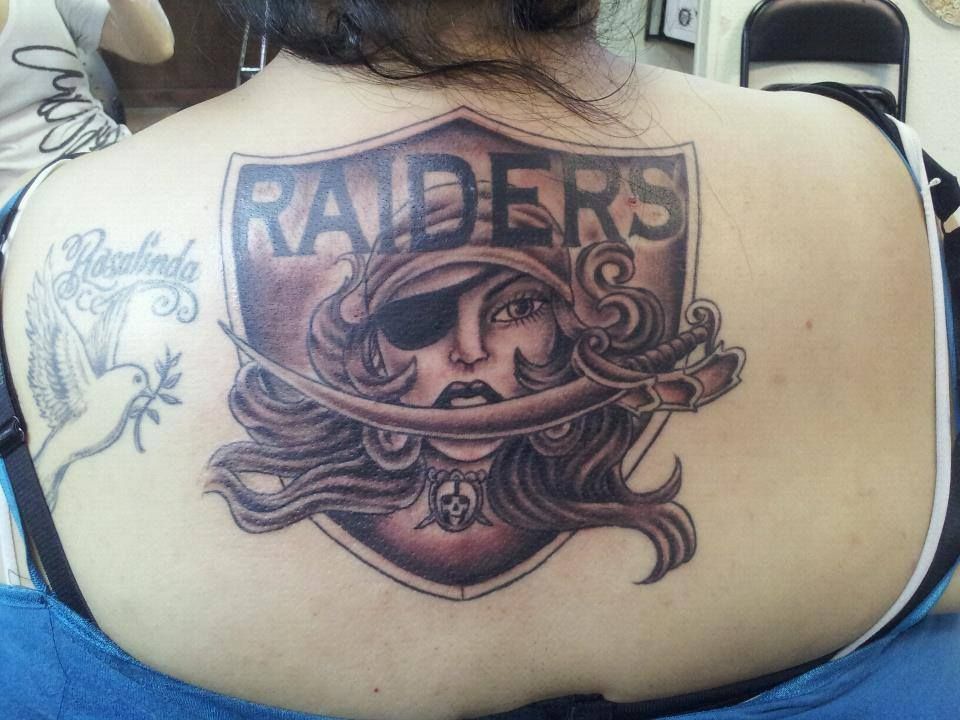 Back tattoo: Raiders shield with pirate woman and sword; other tattoo with dove.