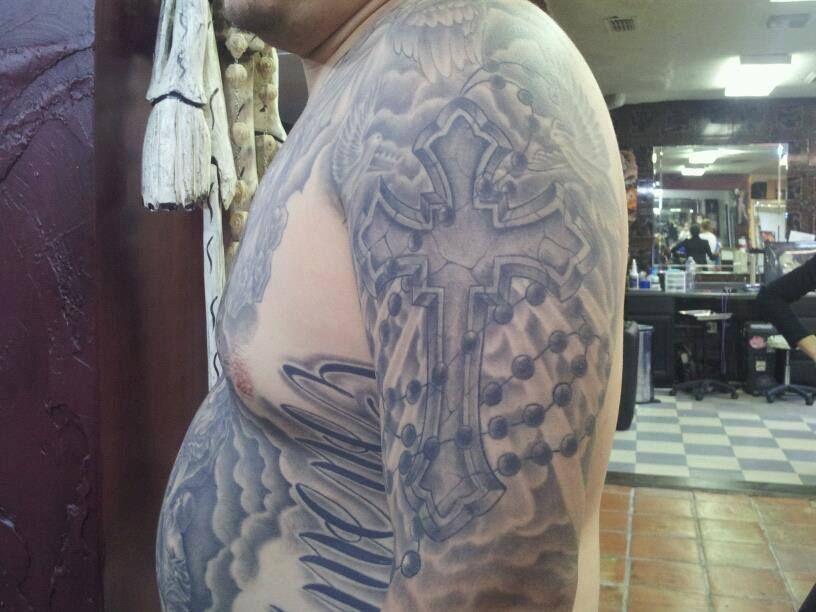 Man's arm with a gray cross and rosary bead tattoo.  Tattoo extends over the shoulder and chest with cloud details.
