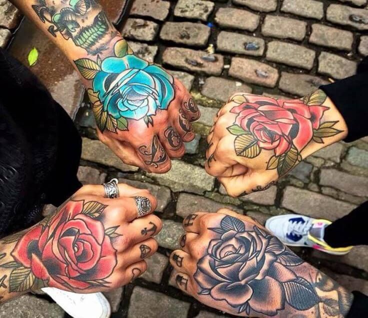 Four hands with rose tattoos in various colors, touching at the knuckles, on cobblestone.