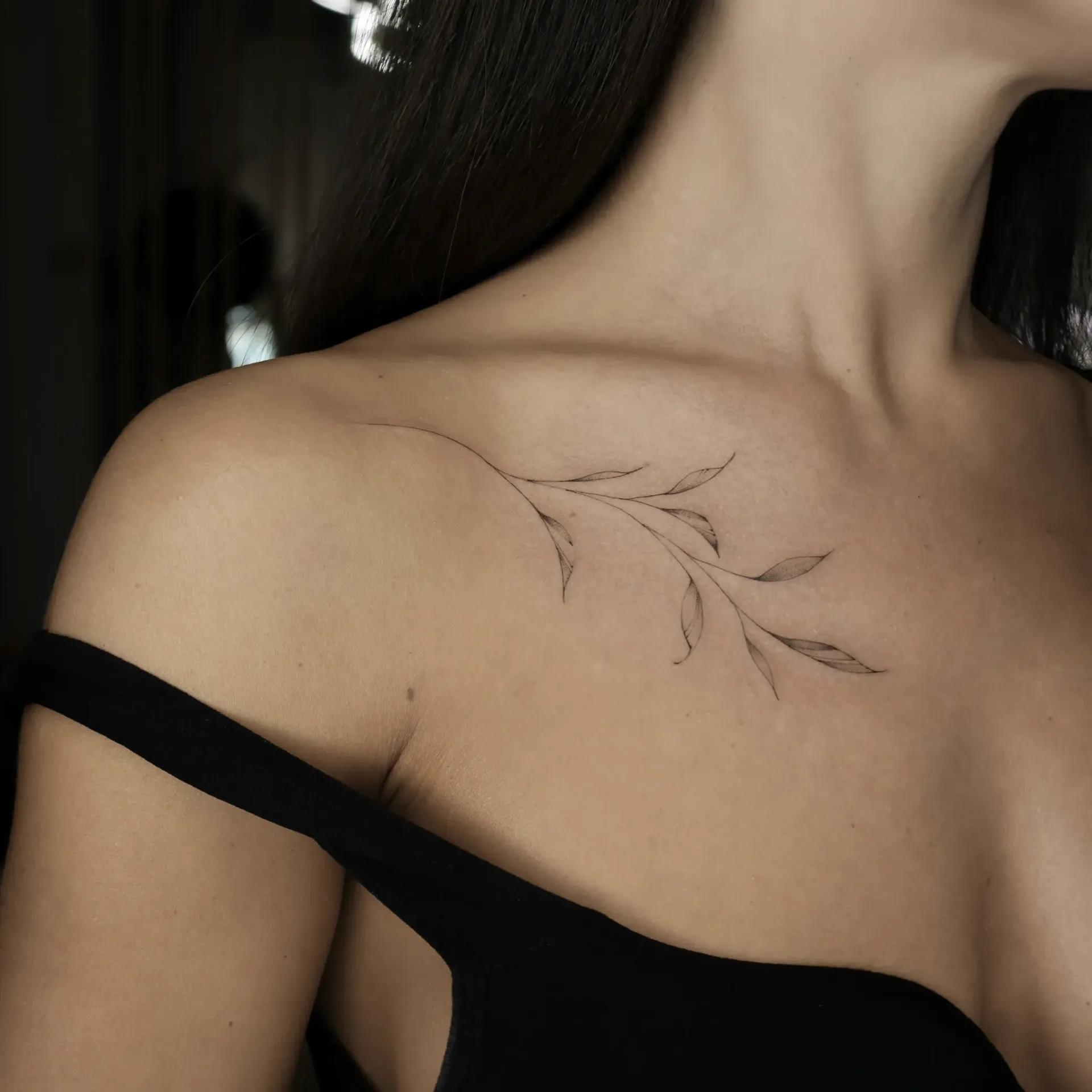 Shoulder of person with black tank top and a tattoo of a thin branch with leaves.