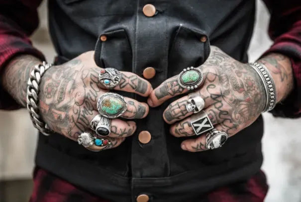 Tattooed hands with rings clasping a black jacket. A silver bracelet and turquoise rings are visible.