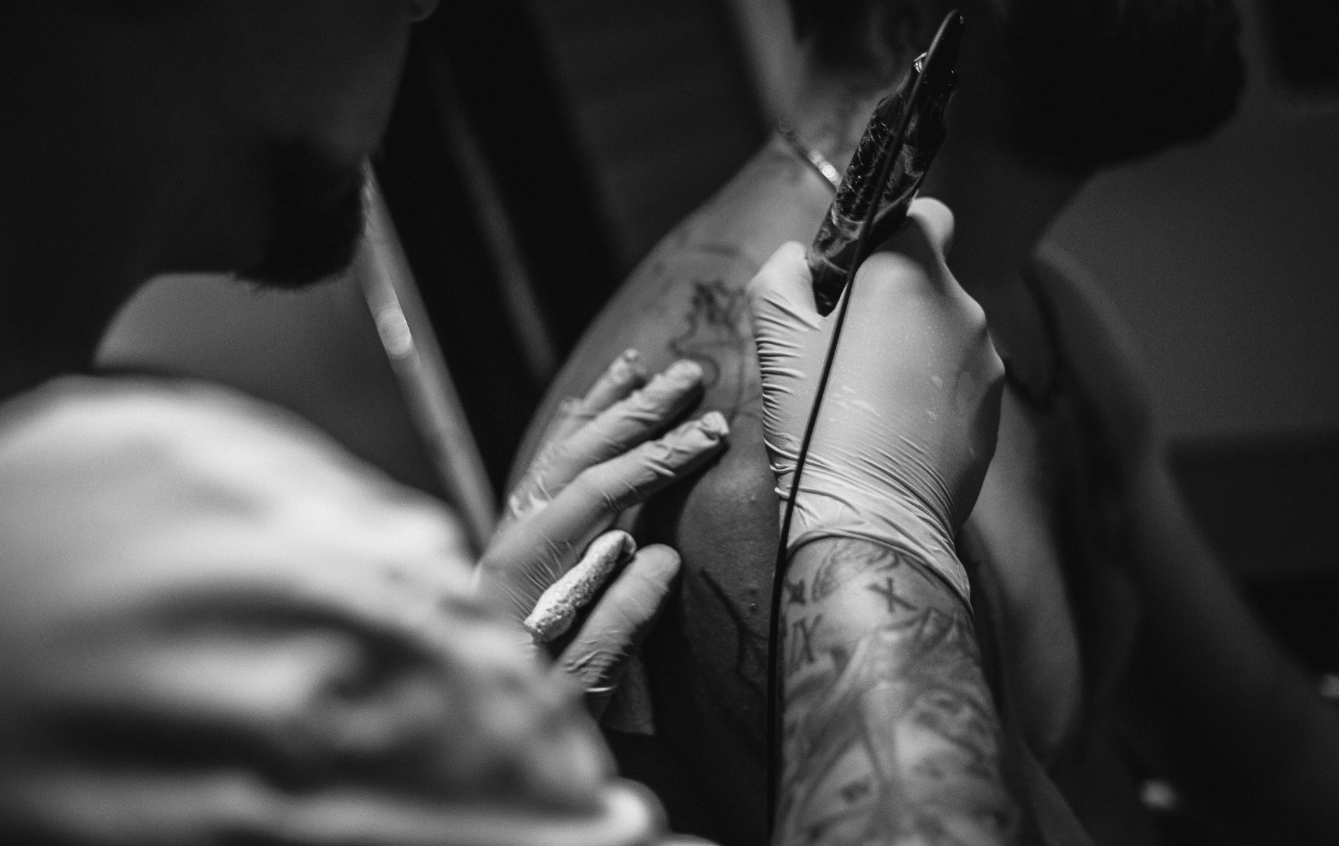 Black and white photo of a tattoo artist giving a shoulder tattoo. The artist uses a machine, gloved hands visible.