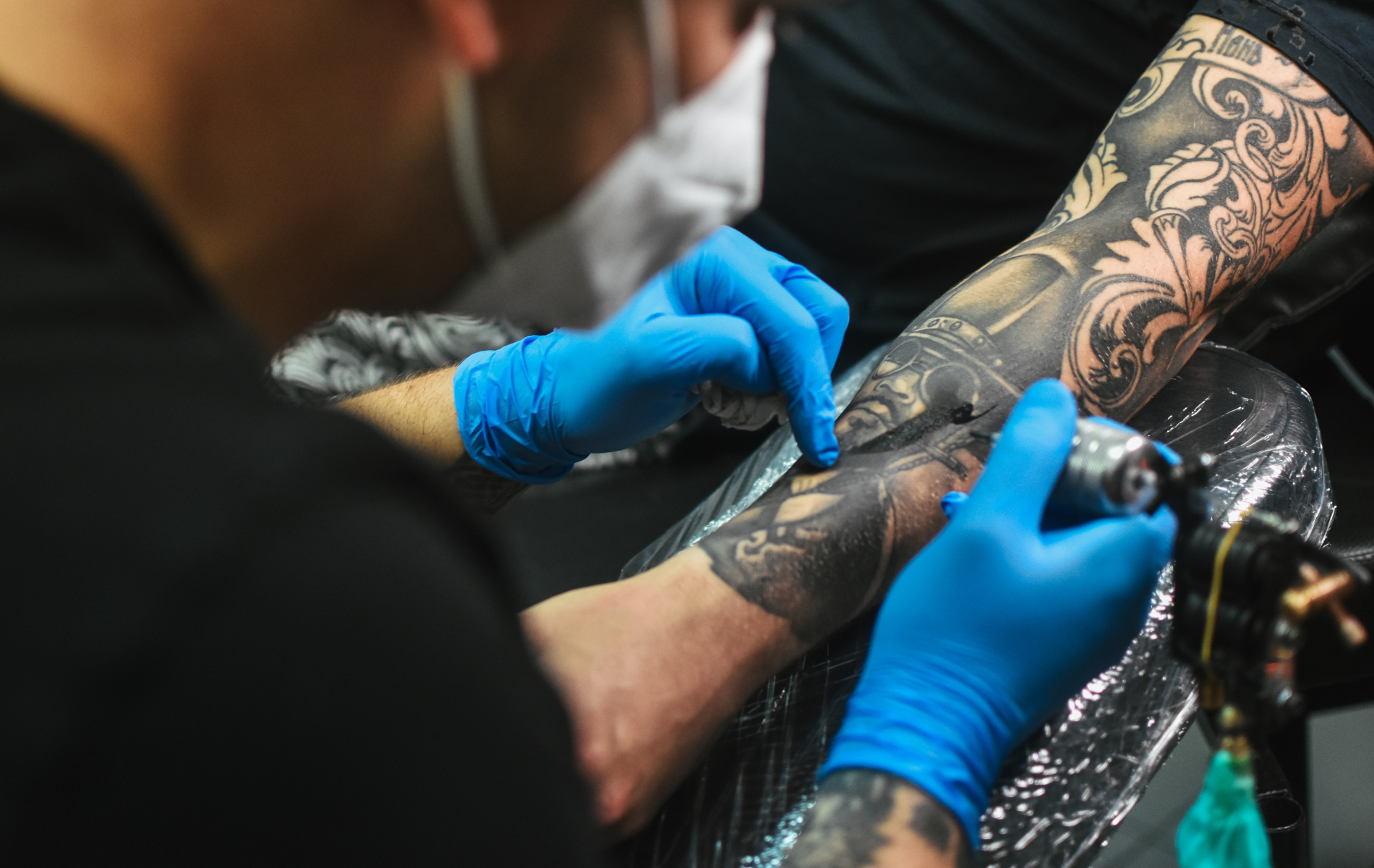 Tattoo artist with blue gloves working on a detailed black and grey arm tattoo.
