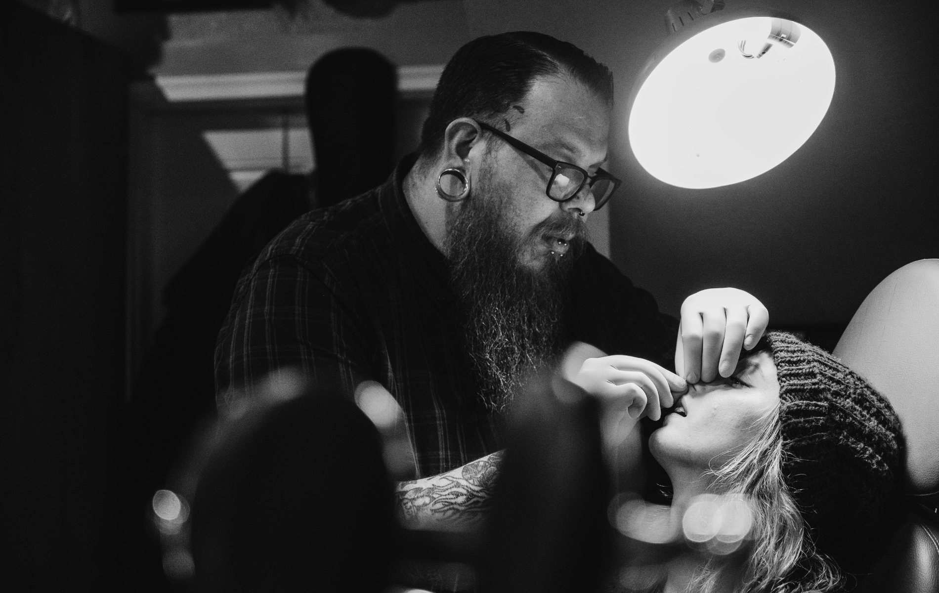 A person with a beard and glasses is performing a nose piercing on another person under a bright light.