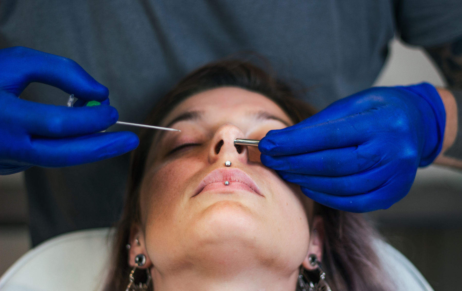 Person getting nose pierced with blue-gloved hands holding tools.