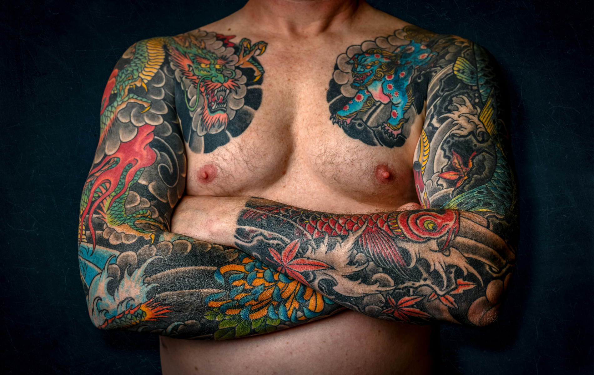 Man with full-body Japanese-style tattoos, arms crossed over chest; dark background.