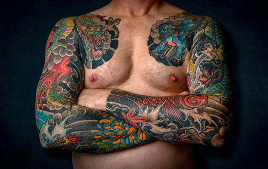 Man with full-body Japanese-style tattoos, arms crossed over chest; dark background.