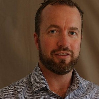 Man with short brown hair and beard, wearing a patterned blue shirt, looking toward the camera.