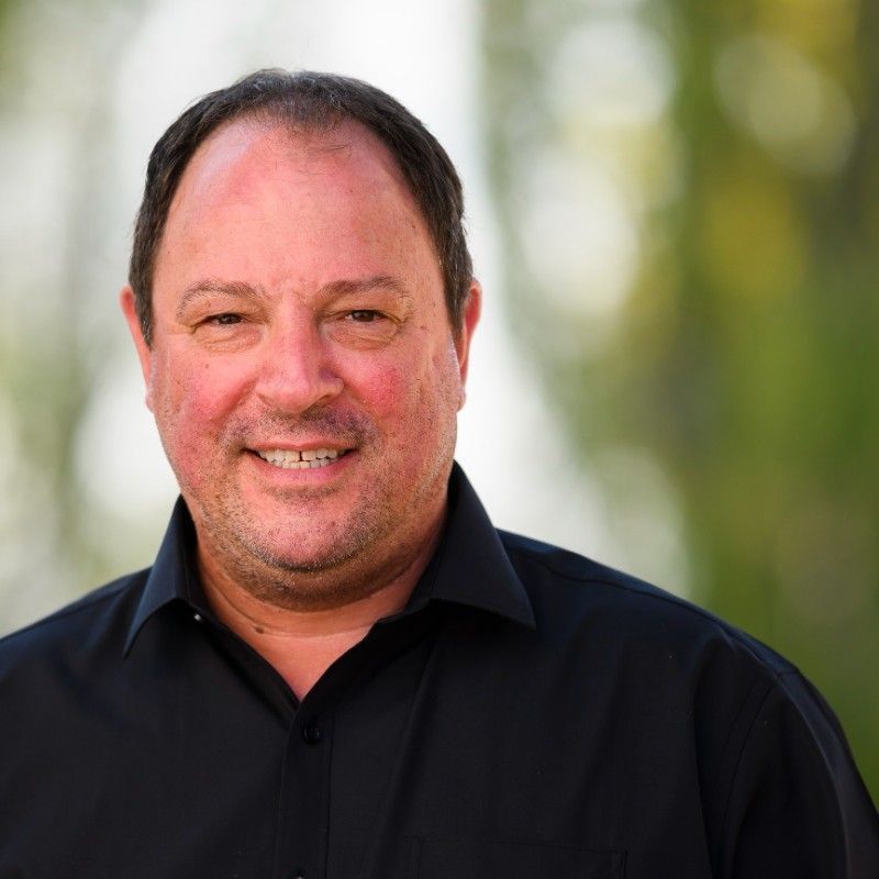 Man with short dark hair, smiling, wearing a black shirt, set outdoors with blurred greenery.