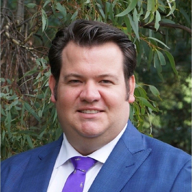 Man in blue suit and purple tie smiles, outdoors with green foliage in background.
