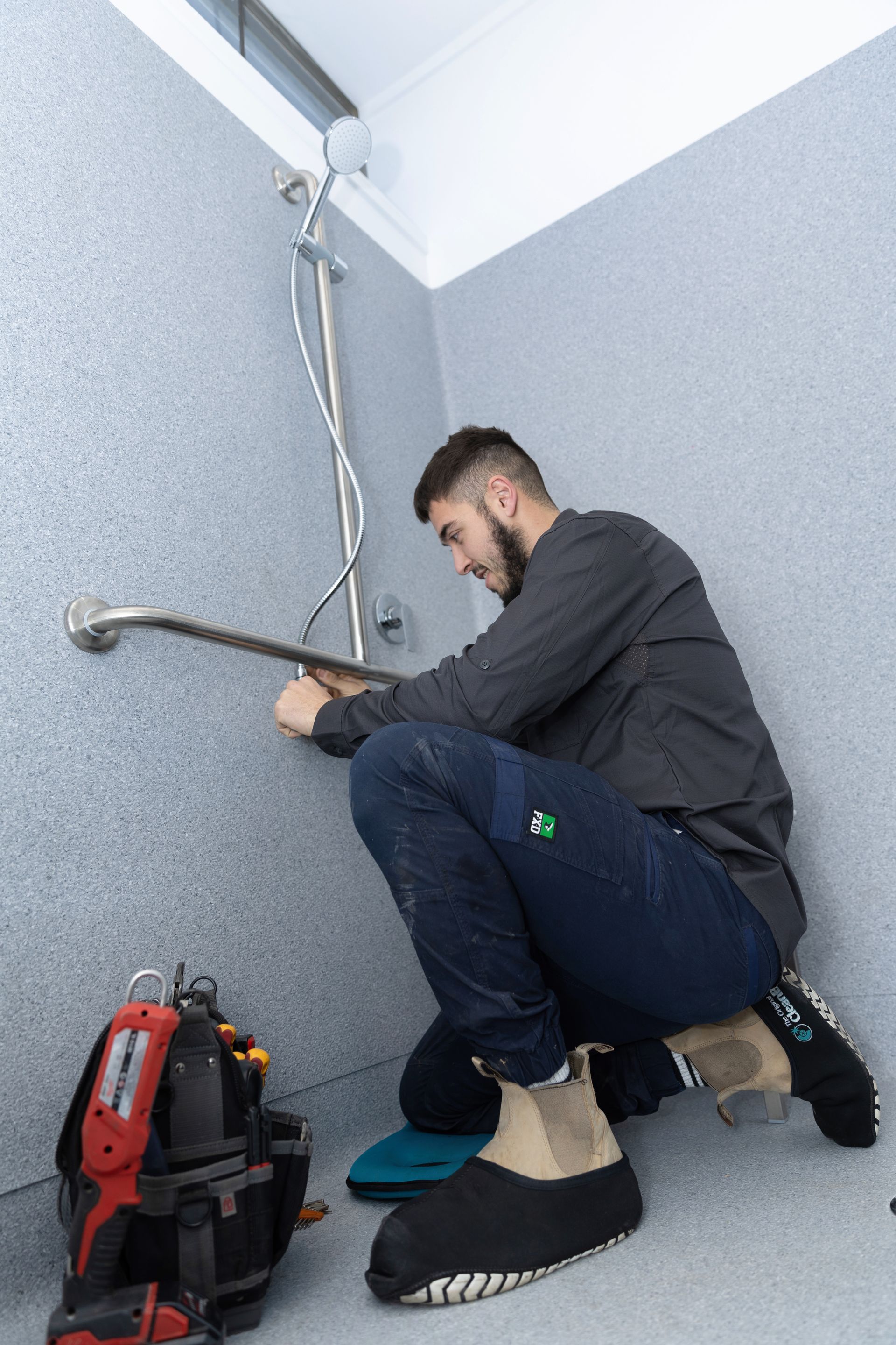 A person installs a grab bar in a shower. They're kneeling, surrounded by tools and grey walls.