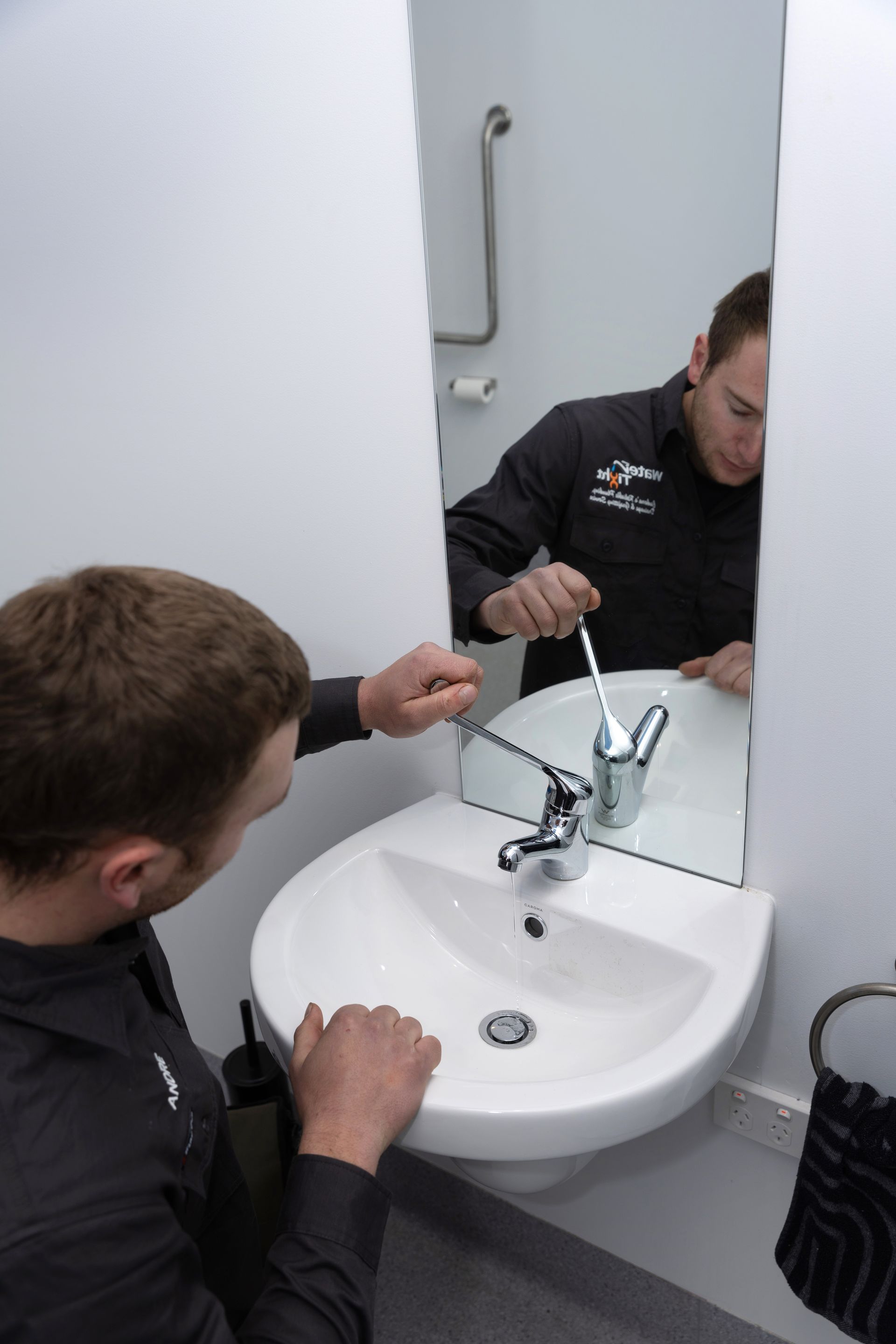 A man in a black shirt is using a wrench on a faucet in front of a mirror in a small, white bathroom.