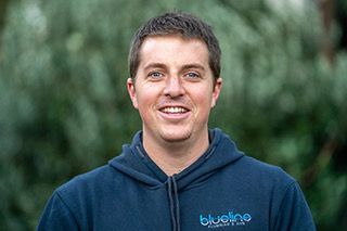 Man in blue hooded sweatshirt smiles outdoors. Blueline Plumbing & Gas logo on the chest.