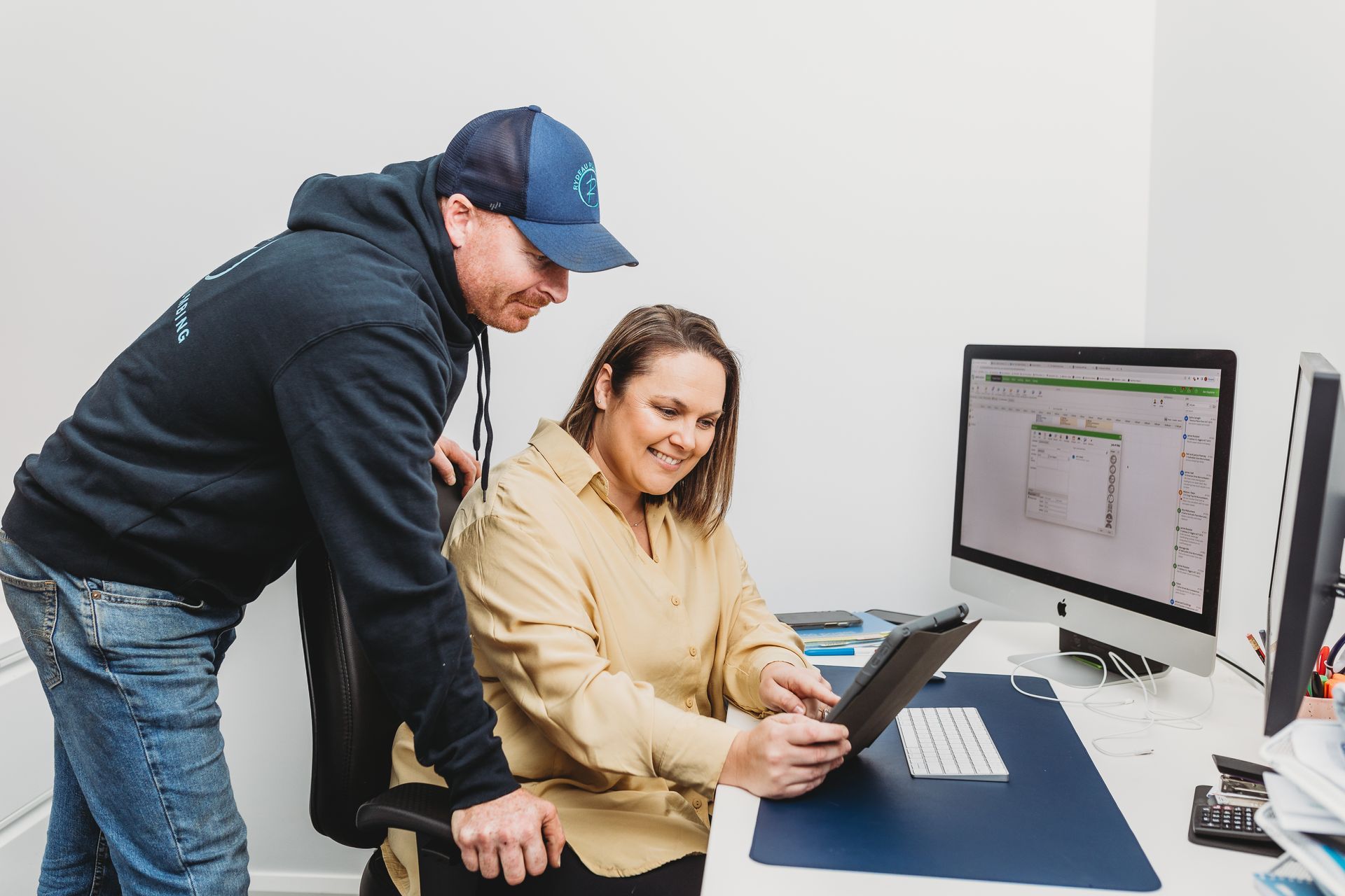 Man in baseball cap and woman looking at a tablet together in an office, smiling. A computer monitor is visible.