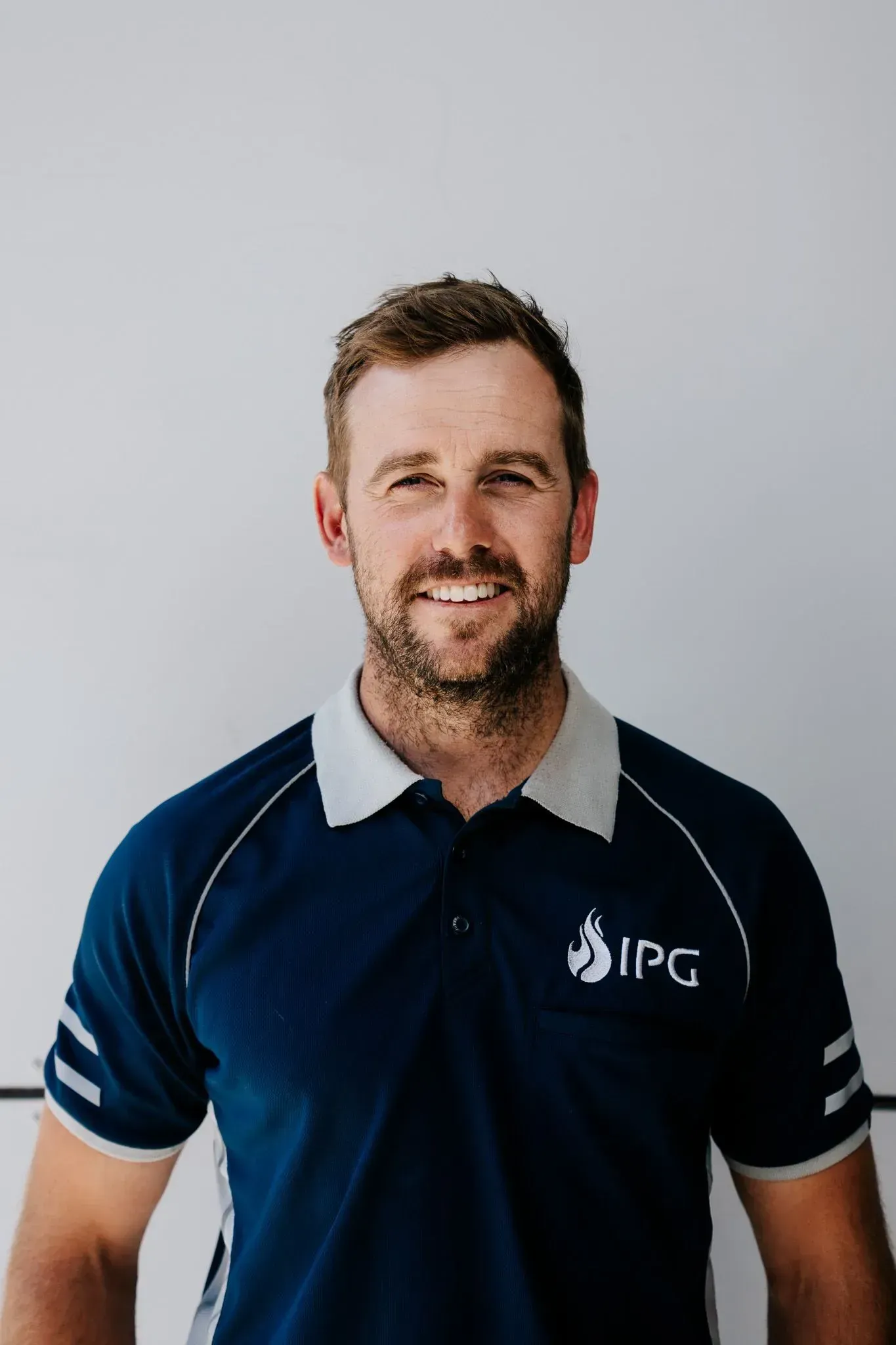 Man with a beard smiling, wearing a navy polo shirt with white trim and IPG logo, standing in front of a white wall.