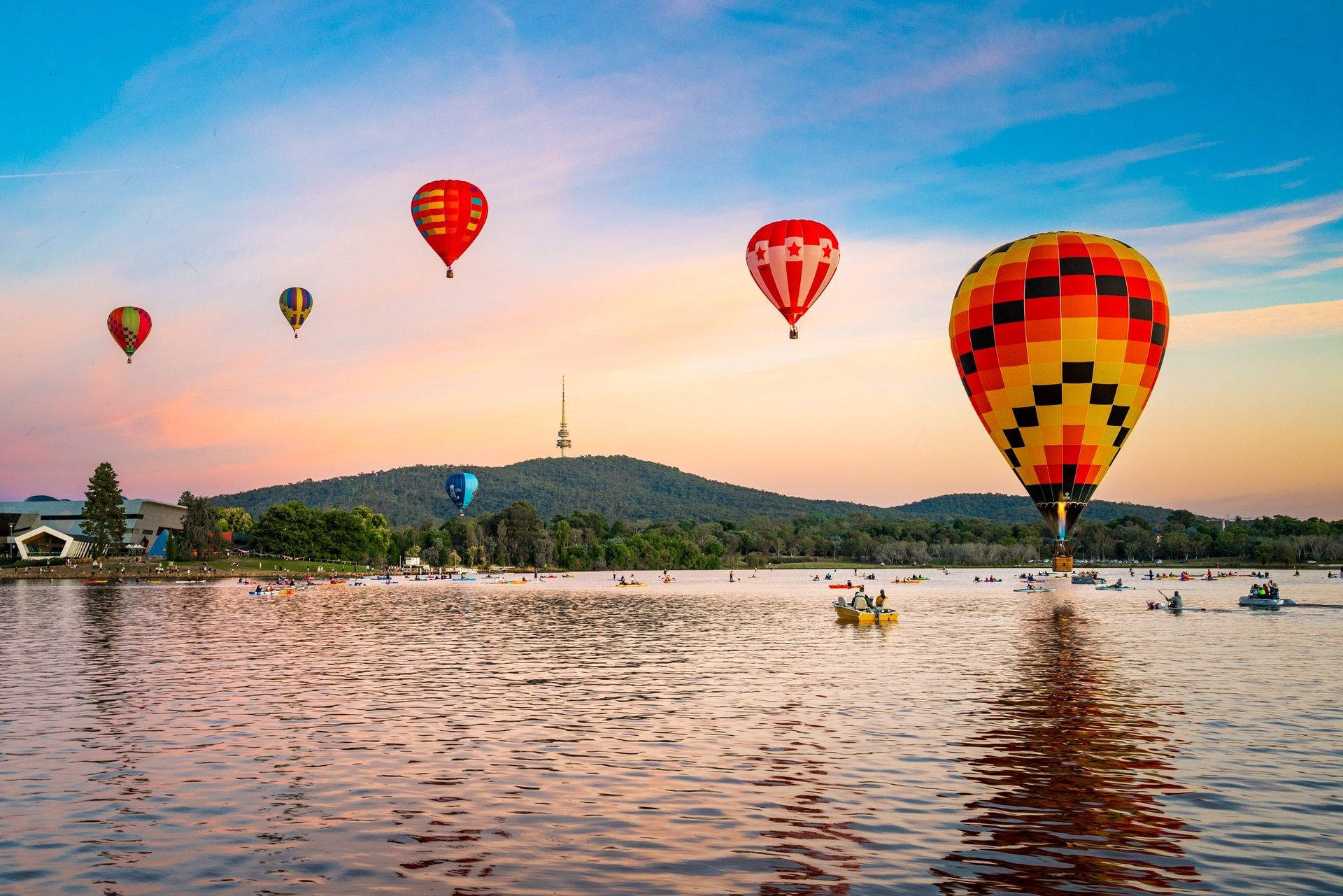 Hot air balloons float over a lake at sunrise, with a mountain and a tower in the distance.