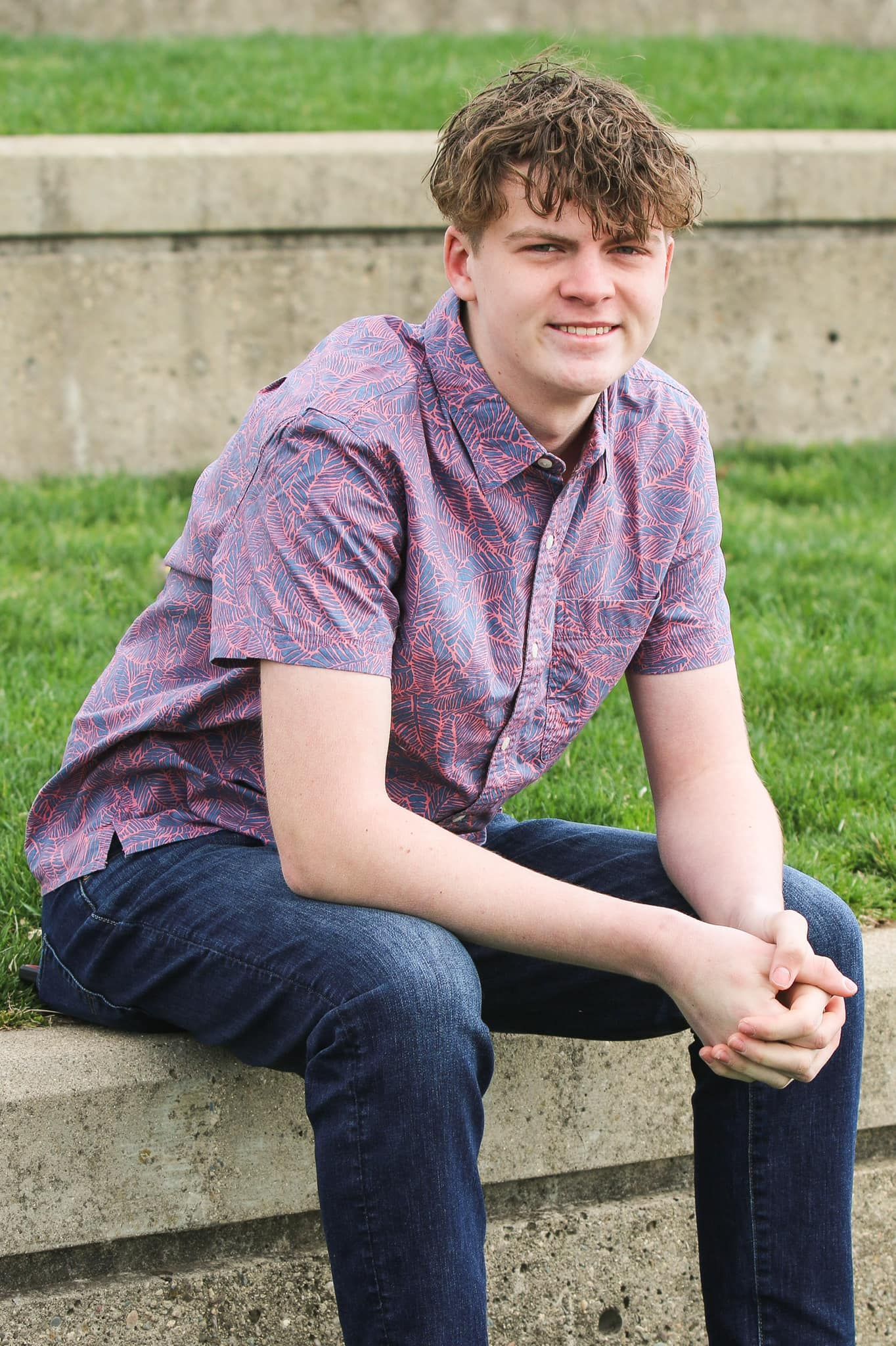A young man is sitting on a stone wall in the grass.