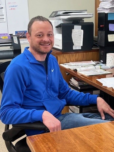 A man in a blue nike jacket sits at a desk