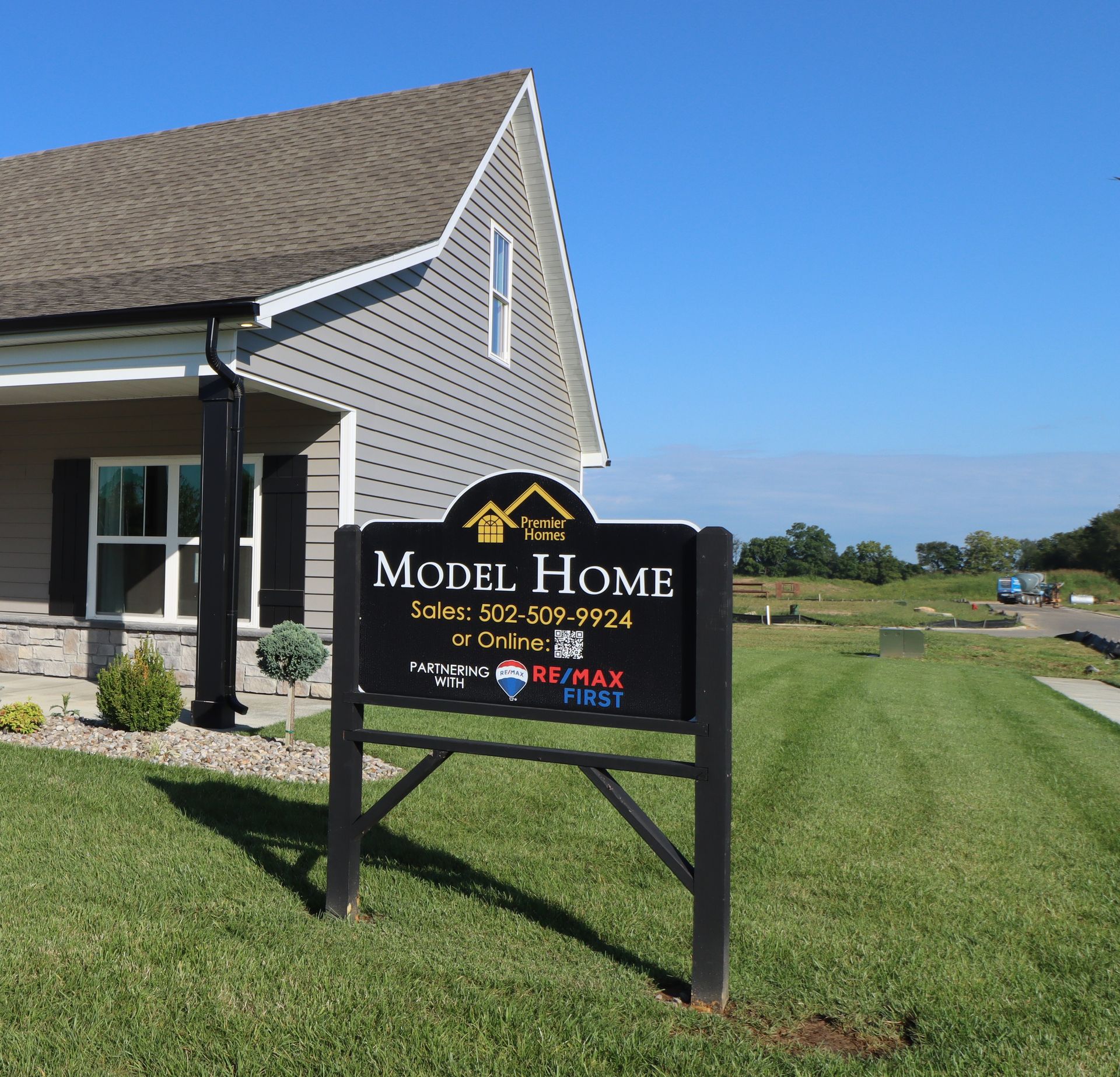 A Model Home sign stands on a manicured lawn in front of a gray-sided house under a clear blue sky.