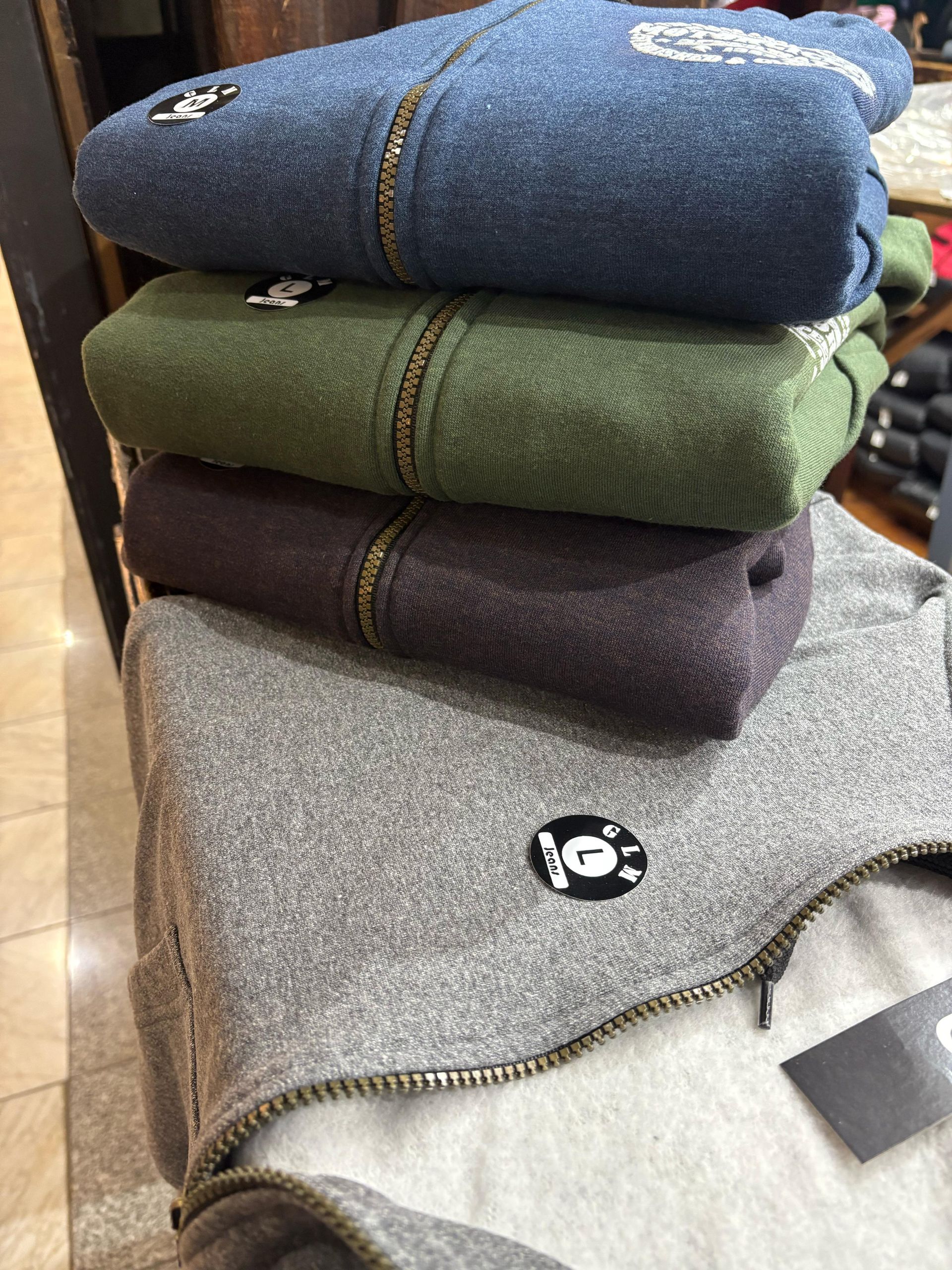 Hombre con sudadera gris con cremallera, manos en los bolsillos, sonriendo. Fondo de pared de madera.