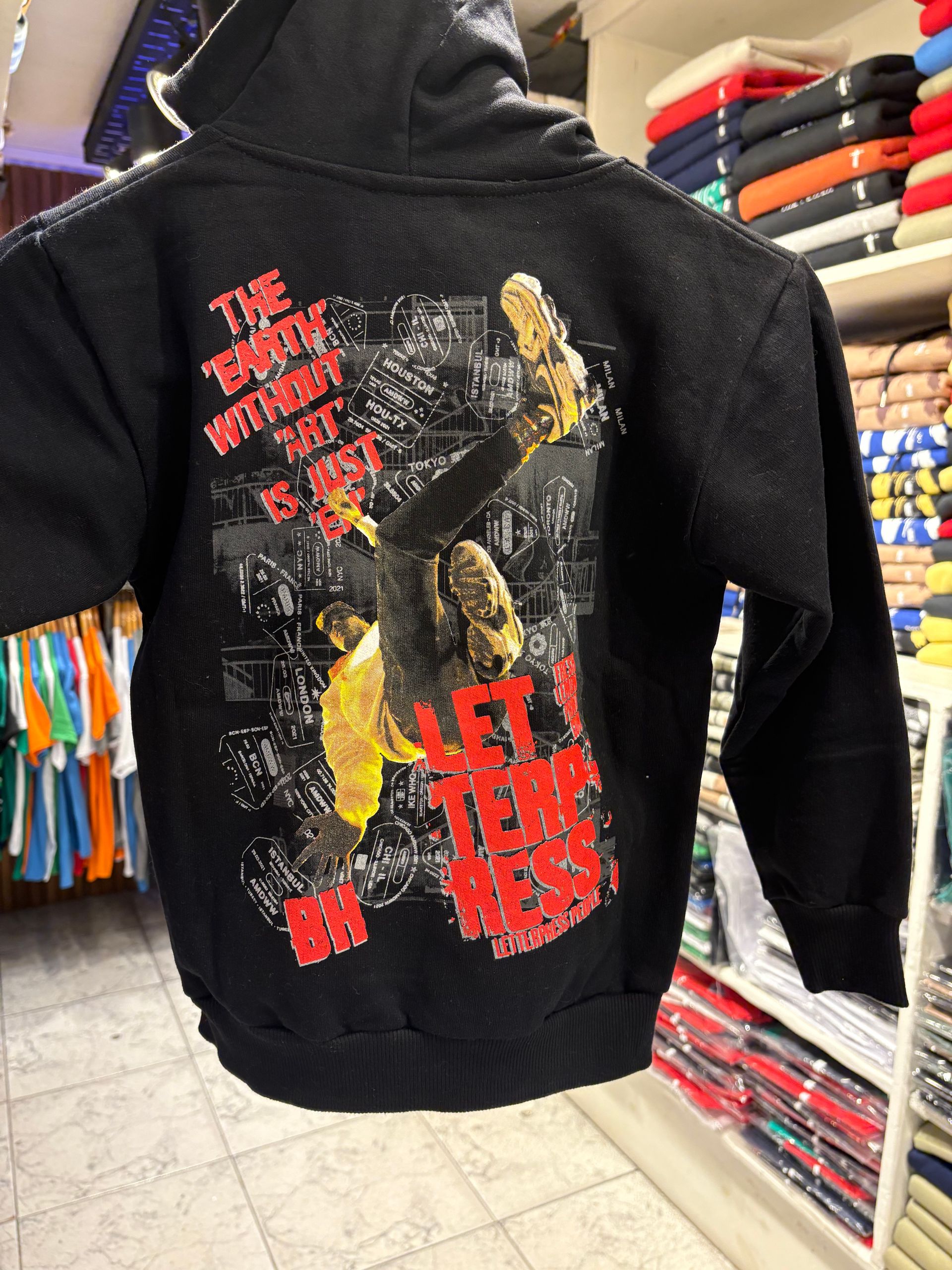 Sudadera con capucha negra con gráfico rojo y etiqueta de precio, colgada en una percha de madera en una tienda.