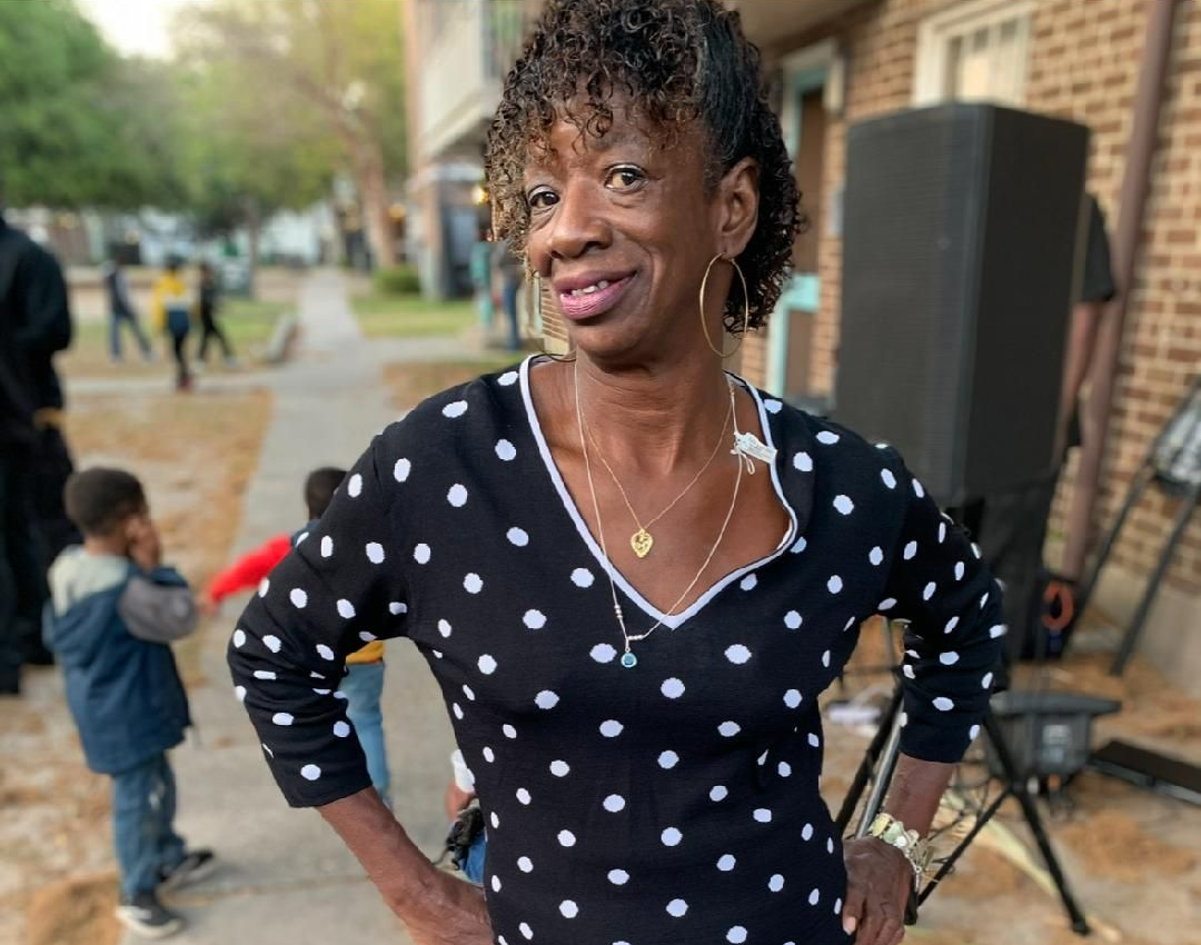 Woman standing on a sidewalk, wearing a black polka-dot top and black pants, smiling with hands on hips