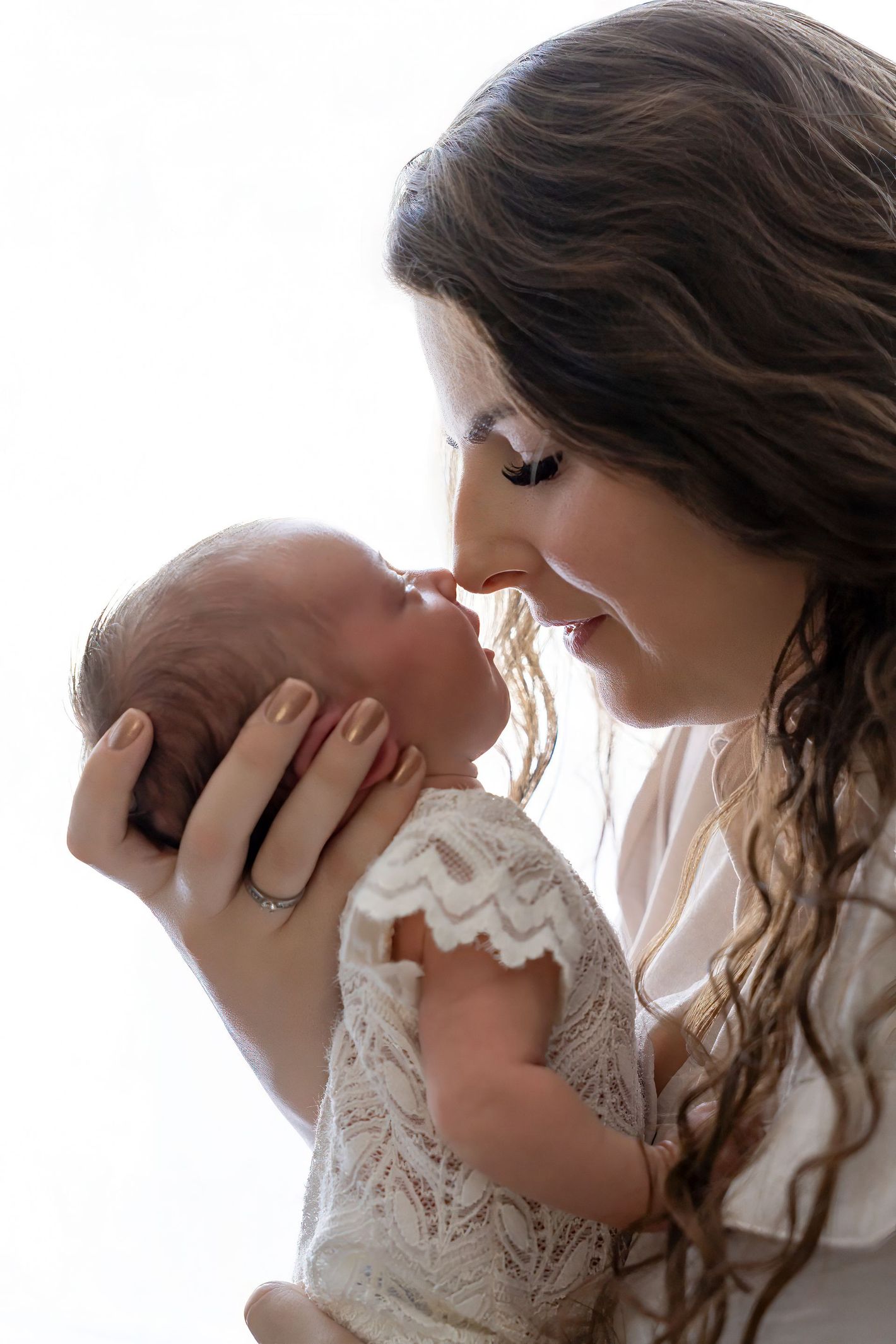 Vrouw raakt zachtjes de neusjes van een baby aan, gekleed in wit kant, tegen een helderwitte achtergrond.