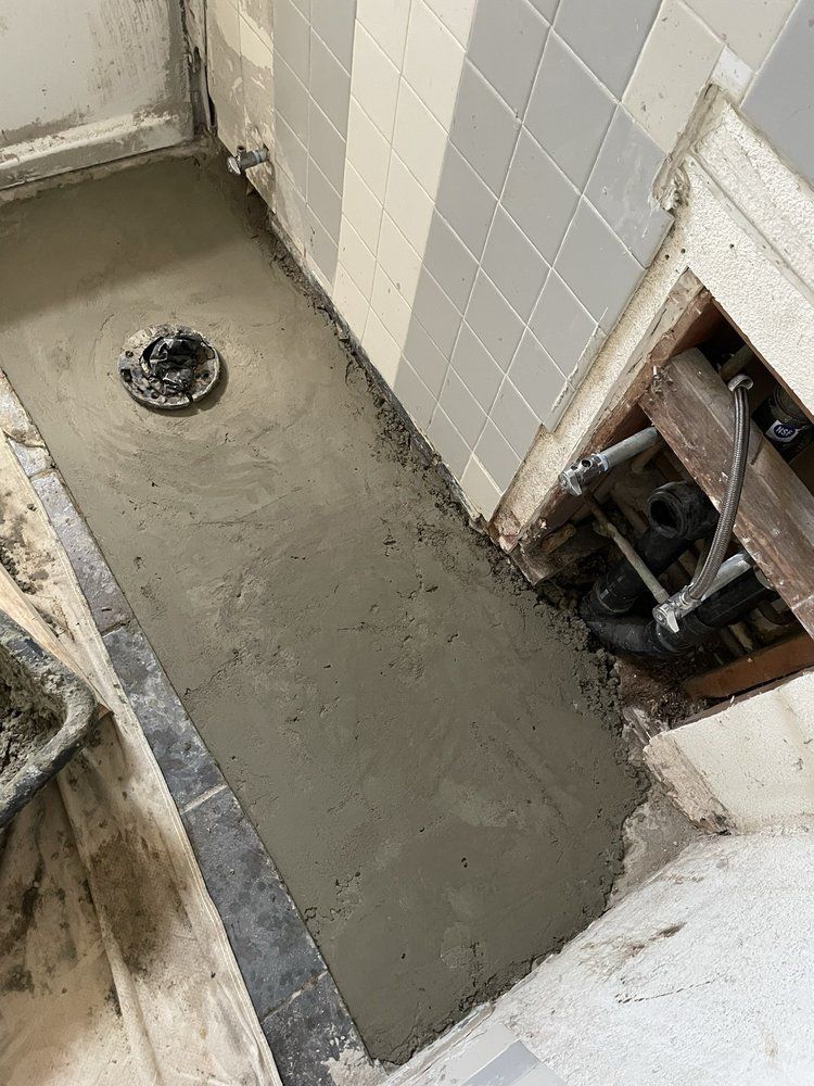 Tiled bathroom floor with a fresh concrete shower base and exposed plumbing on the right.