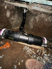 Black PVC pipe repair joint in a dirt crawlspace beneath a floor, with a boot visible nearby.